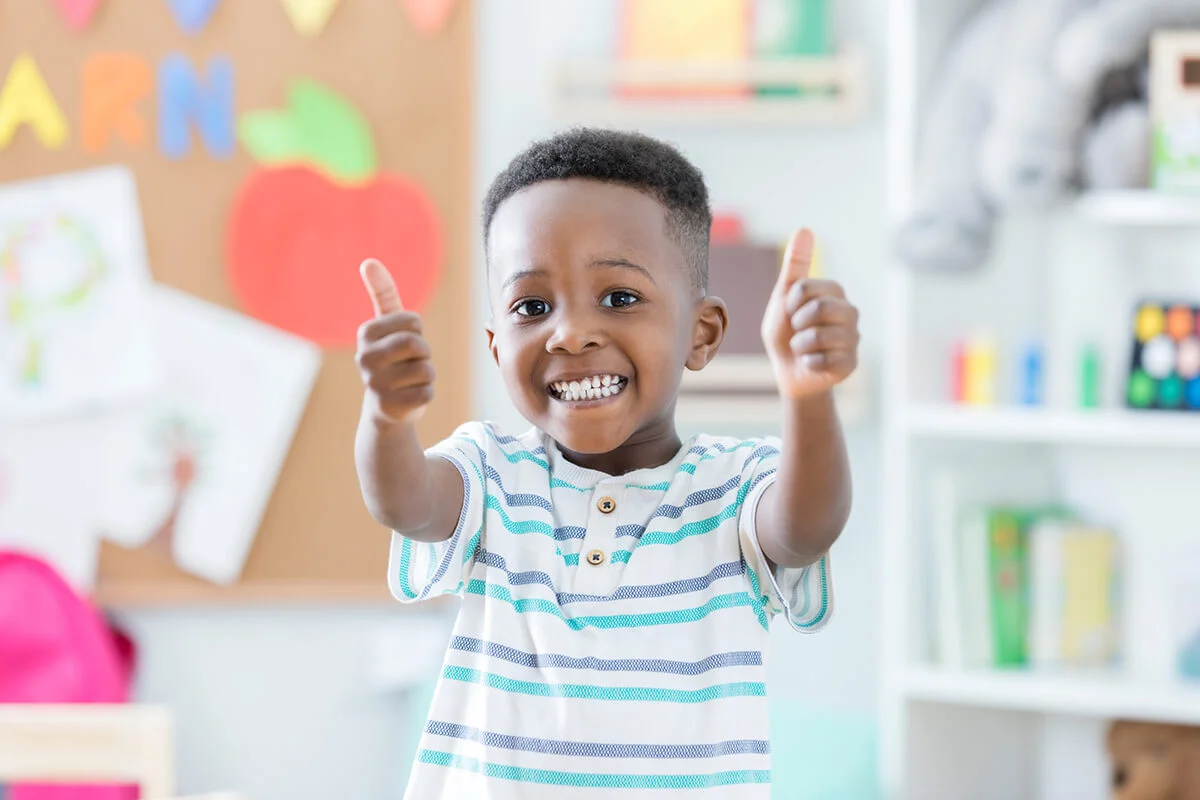 boy smiles for the camera as he stands in his preschool classroom