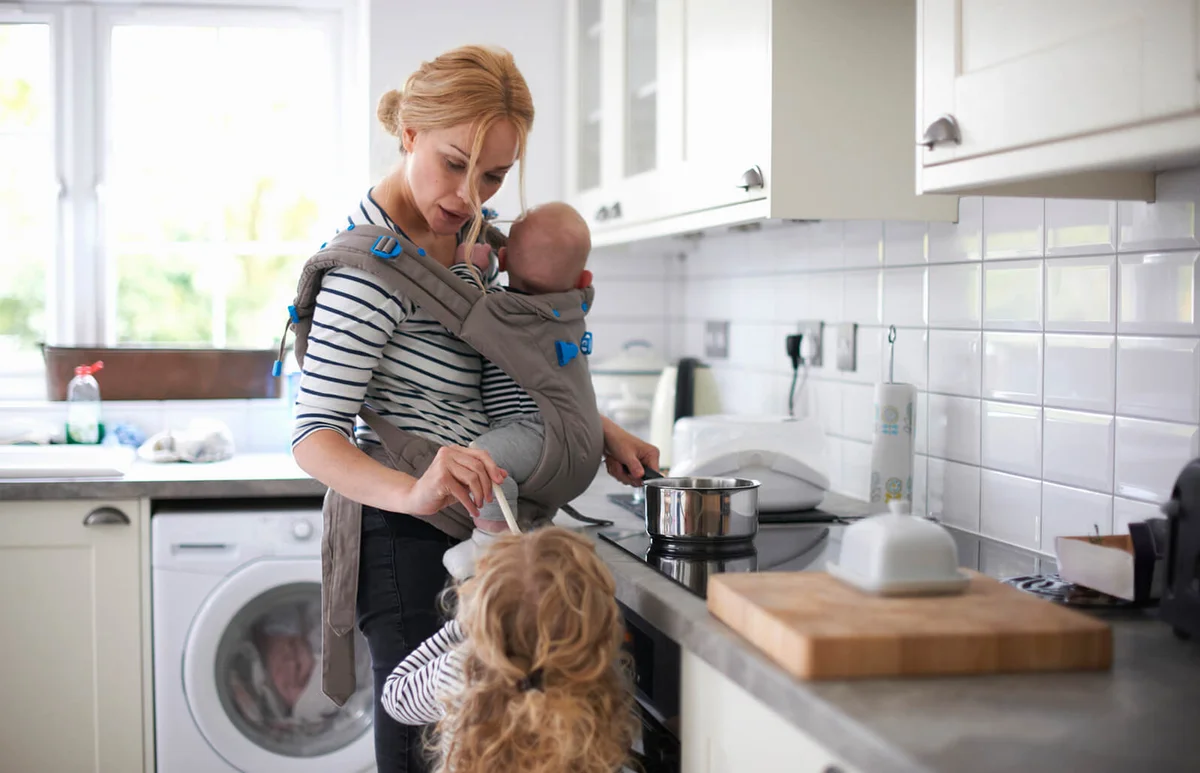 Woman cooking in kitchen, baby strapped to body in sling, daughter standing beside her