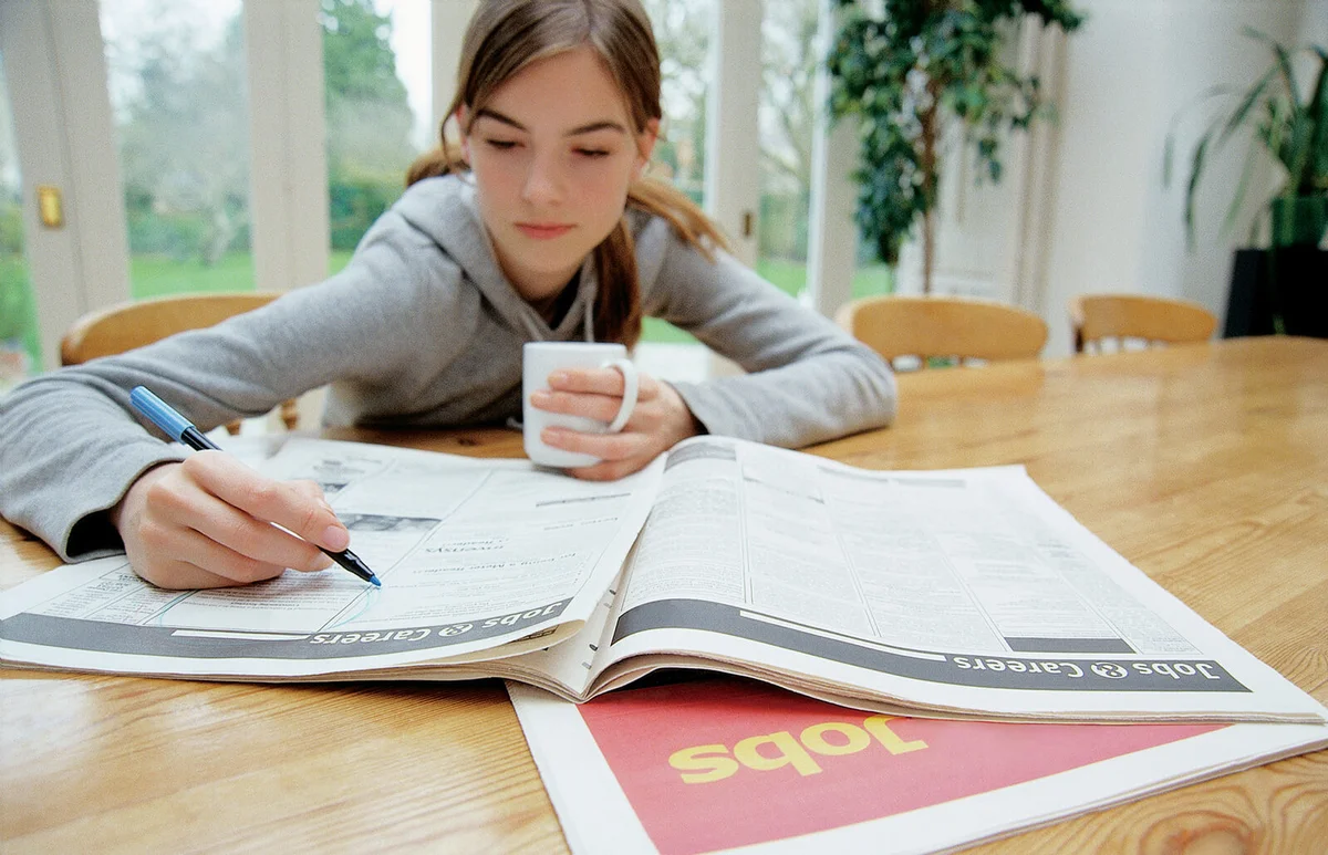 Teenage Girl Sitting Behind a Table Searching for Jobs in a Newspaper