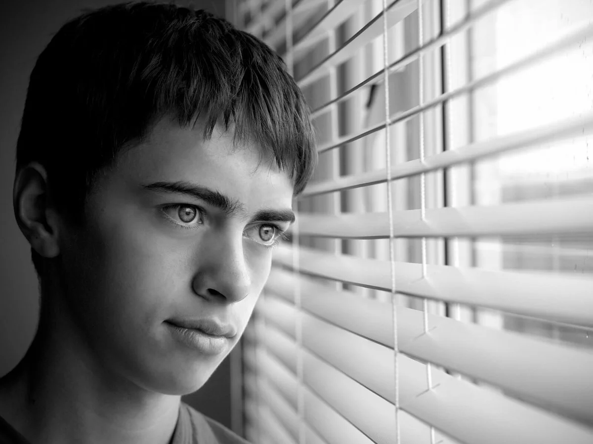Bored young boy looking through window blinds rainy day
