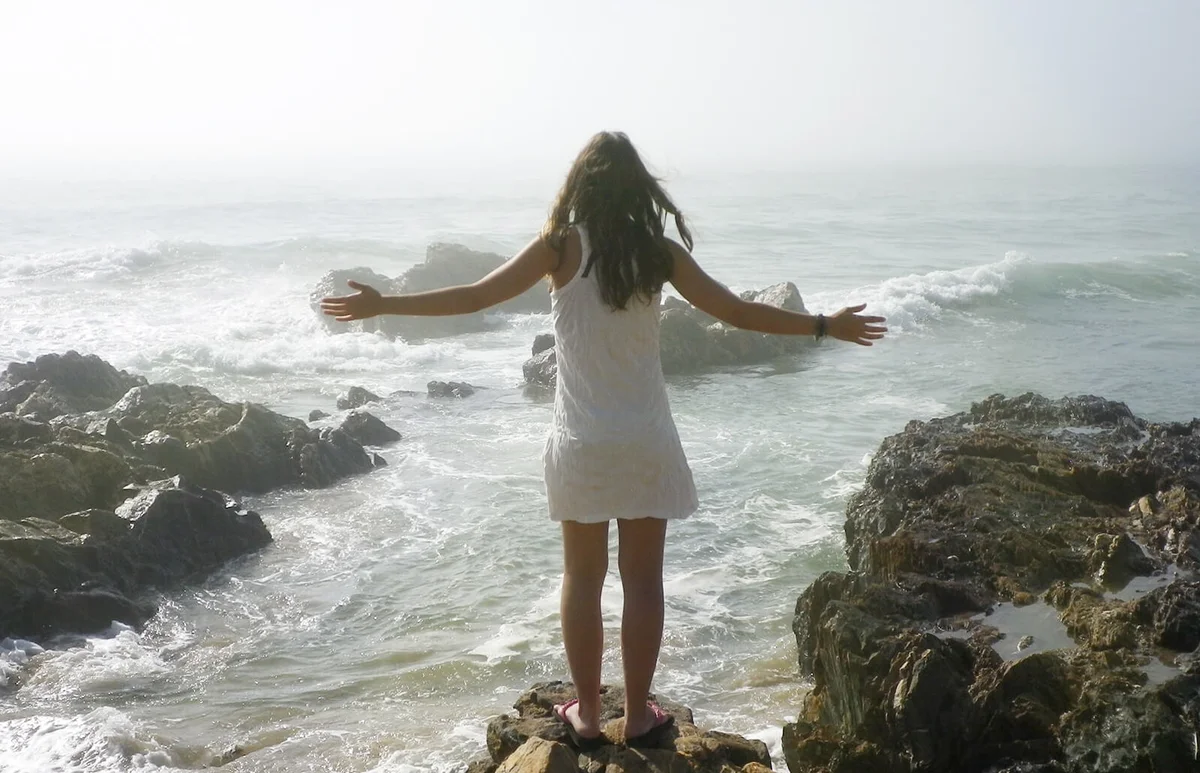 Woman With Arms Outstretched Standing On Rocky Shore