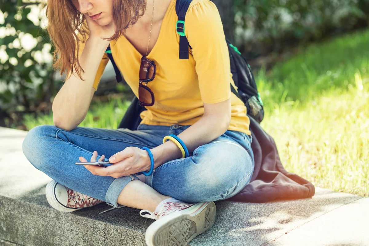 Sad teenage girl sitting alone in the park and reading on her phone