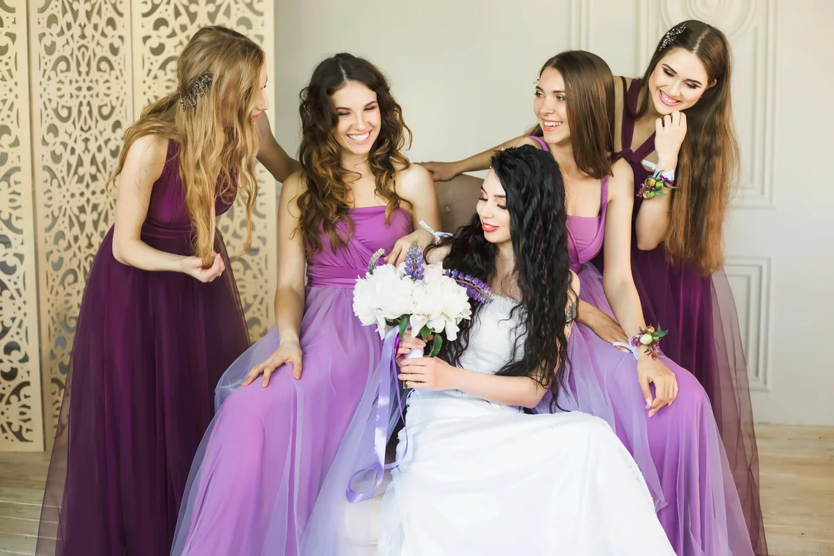 Bride holding a wedding bouquet and smiling while talking to her bridesmaids in purple dresses