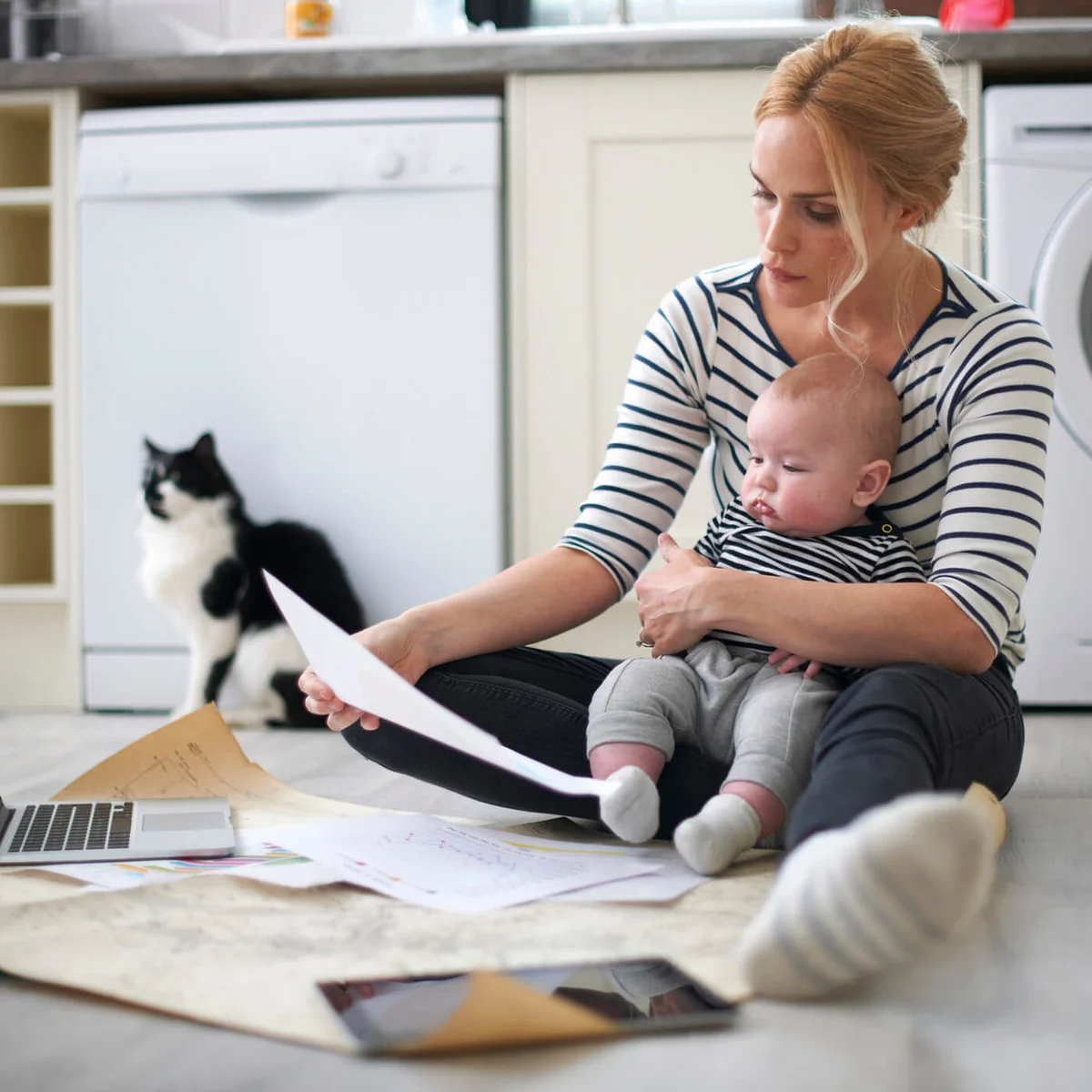 Woman holding baby son in arms, looking through work on kitchen floor