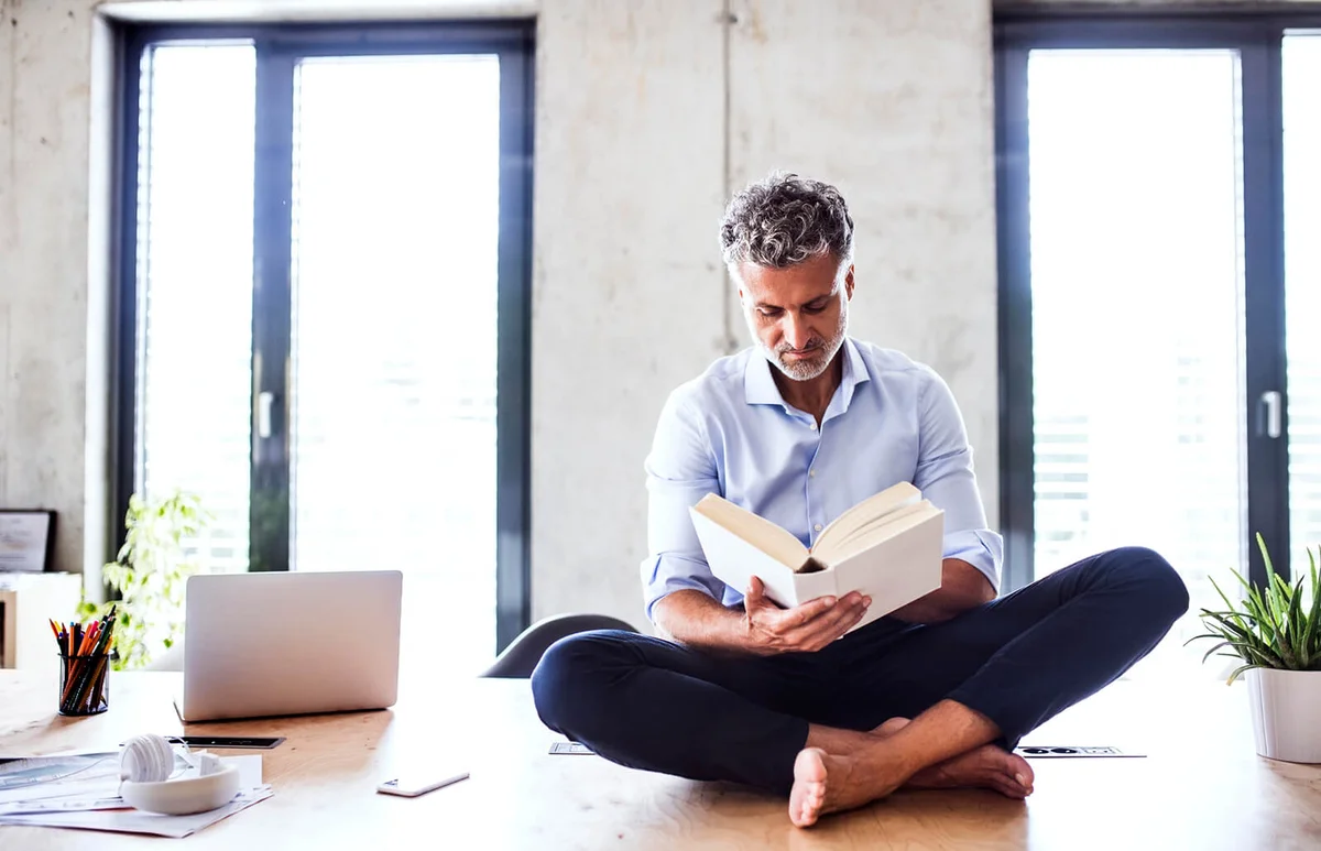 Mature businessman sitting barefoot on desk in office reading a book