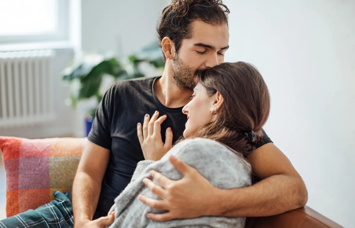 Man embracing girlfriend while kissing on her forehead at home