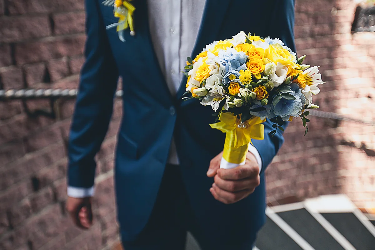 groom in an elegant blue suit is holding a beautiful bouquet of blue, yellow and ivory flowers