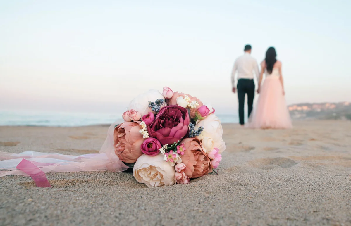 Close-Up Of Bouquet On Sand With Couple In Background