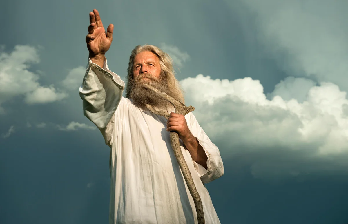 man with long grey hair and a long grey beard standing in front of a dramatic dark sky