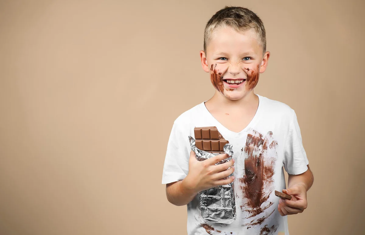 Little boy holds a bar of chocolate in his grin and has smudged clothes