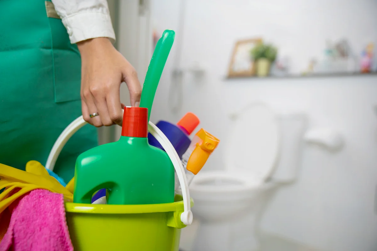 Woman is standing in the bathroom holding a bucket full of cleaning supplies