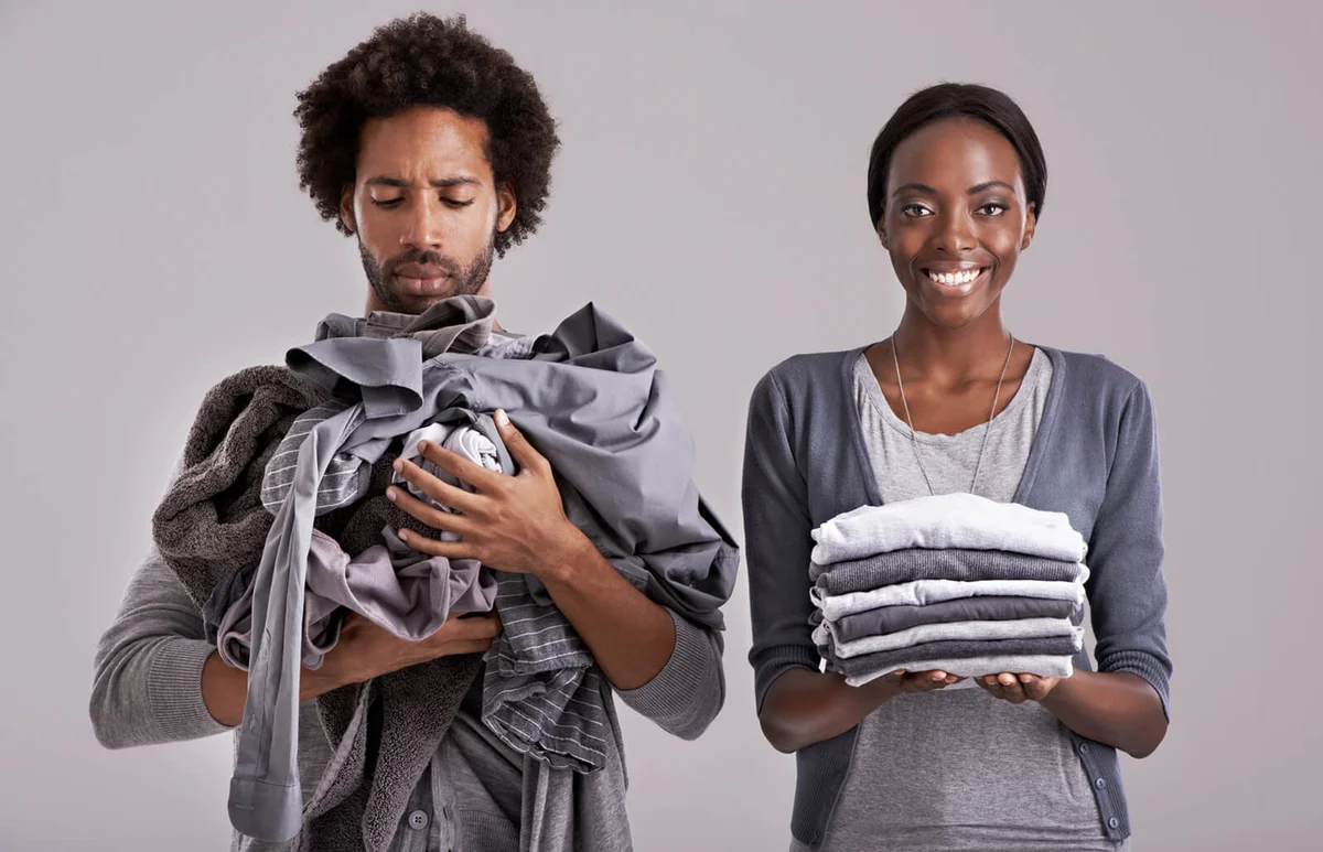 woman holding a neatly folded pile of clothes while her partner is in a mess
