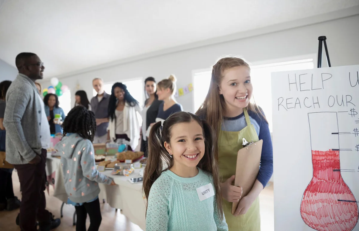 confident girls standing at fundraising poster at bake sale in community center