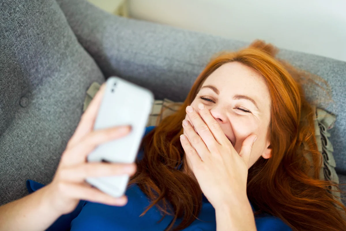 Woman lying on sofa using smart phone, smiling behind hand