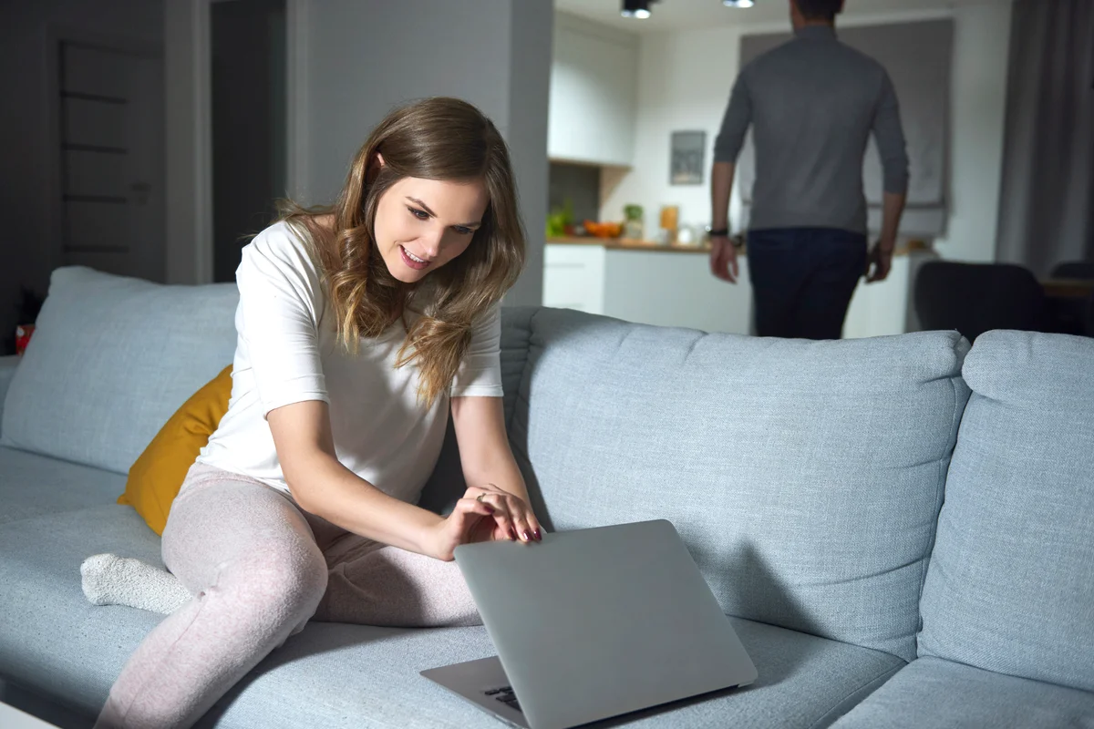 Young woman sitting on sofa in evening, peeking at boyfriend's laptop