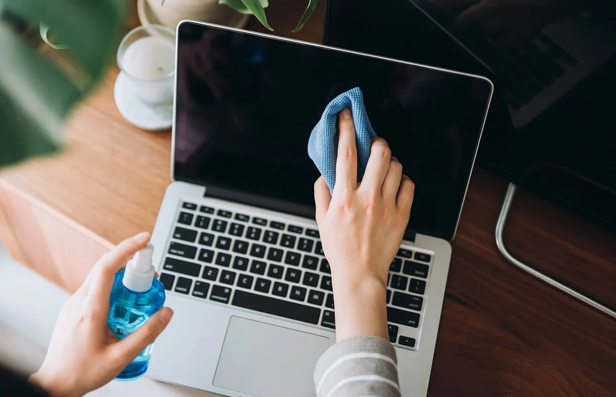young woman cleaning the surface of laptop with cleaning spray and antistatic cloth at home