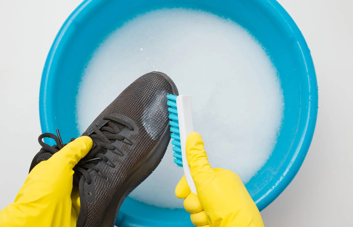 Hands in rubber protective gloves holding black sport shoe and brush above blue basin