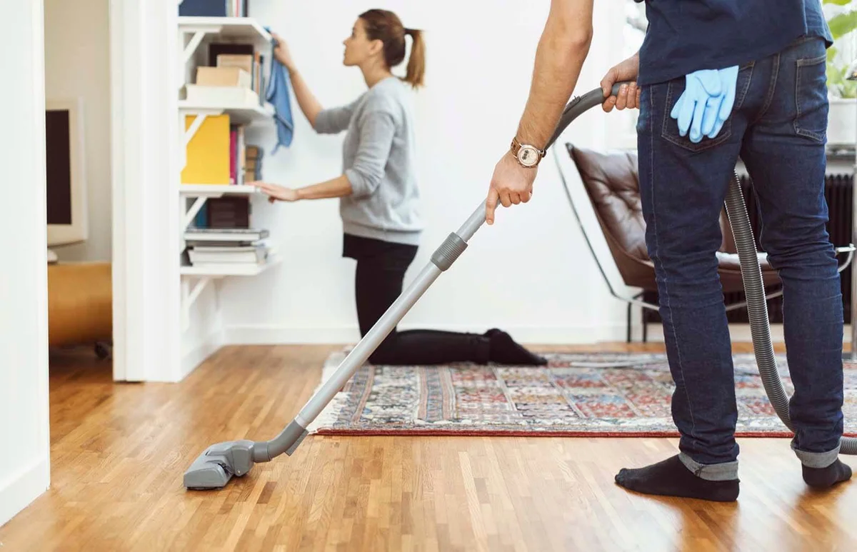 man vacuuming floor while woman cleaning shelves in background at home