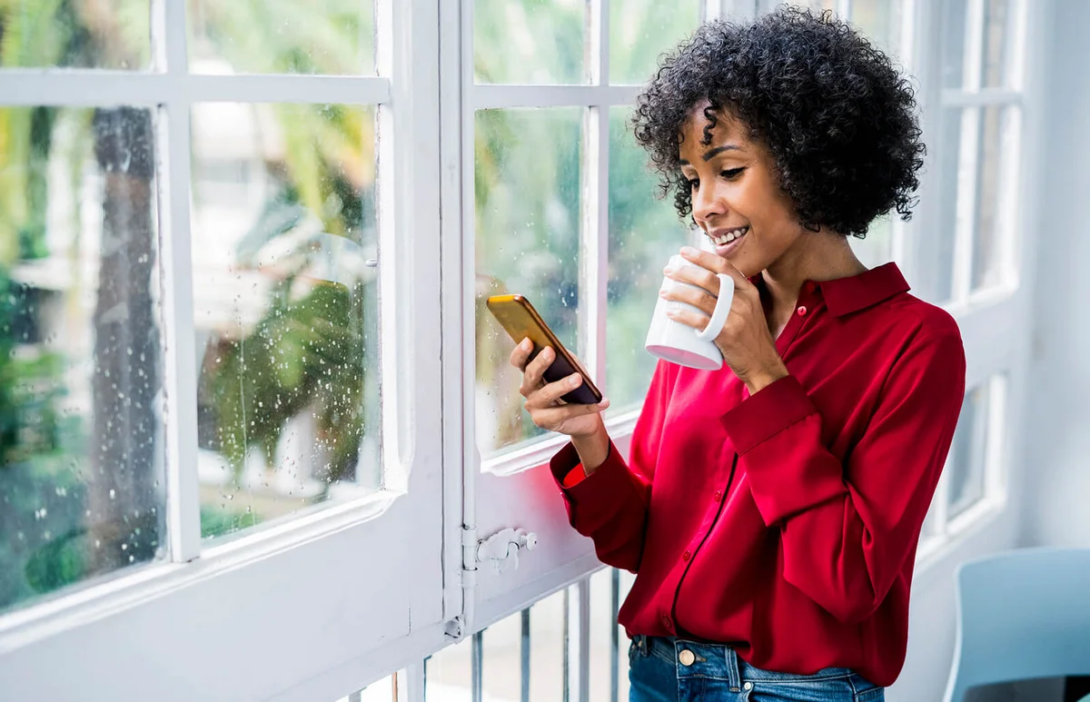 woman with cup of coffee and cell phone standing at the window at home