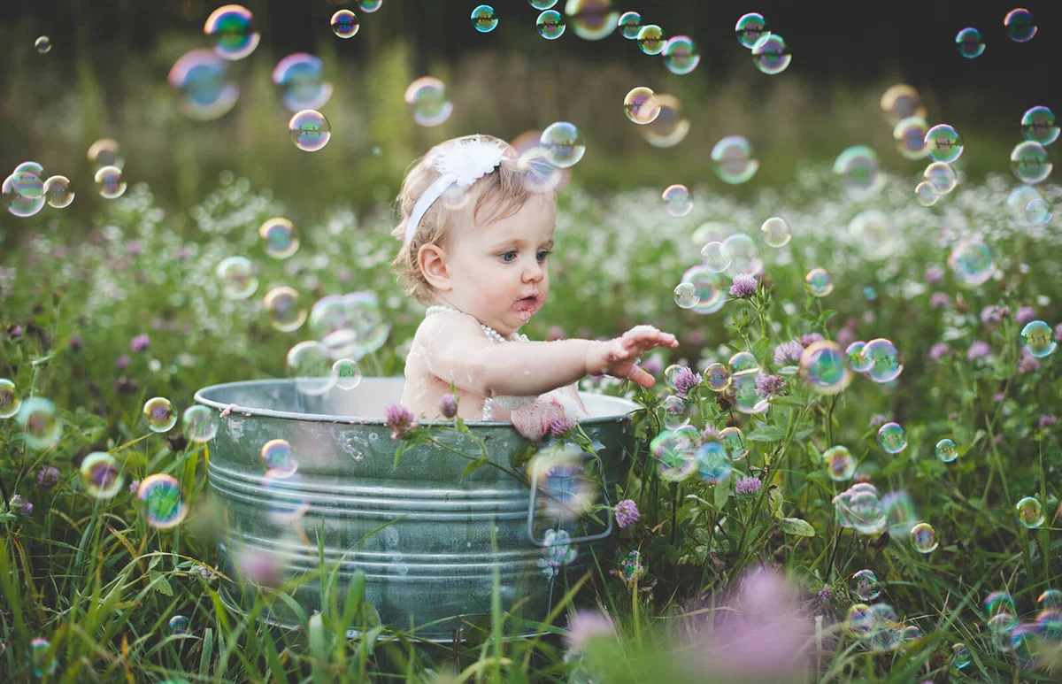 Baby girl in tin bathtub in meadow reaching for floating bubbles