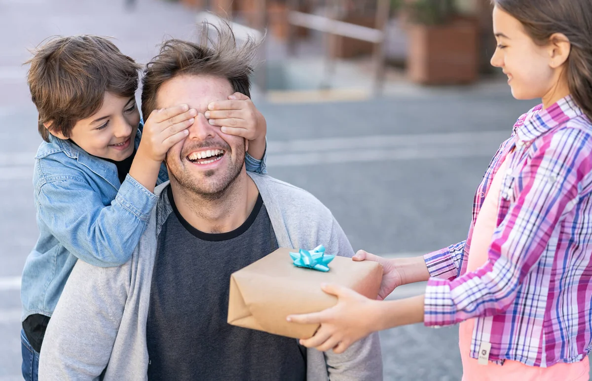 Cute soon covering his father's eyes while daughter holds a present for father's day