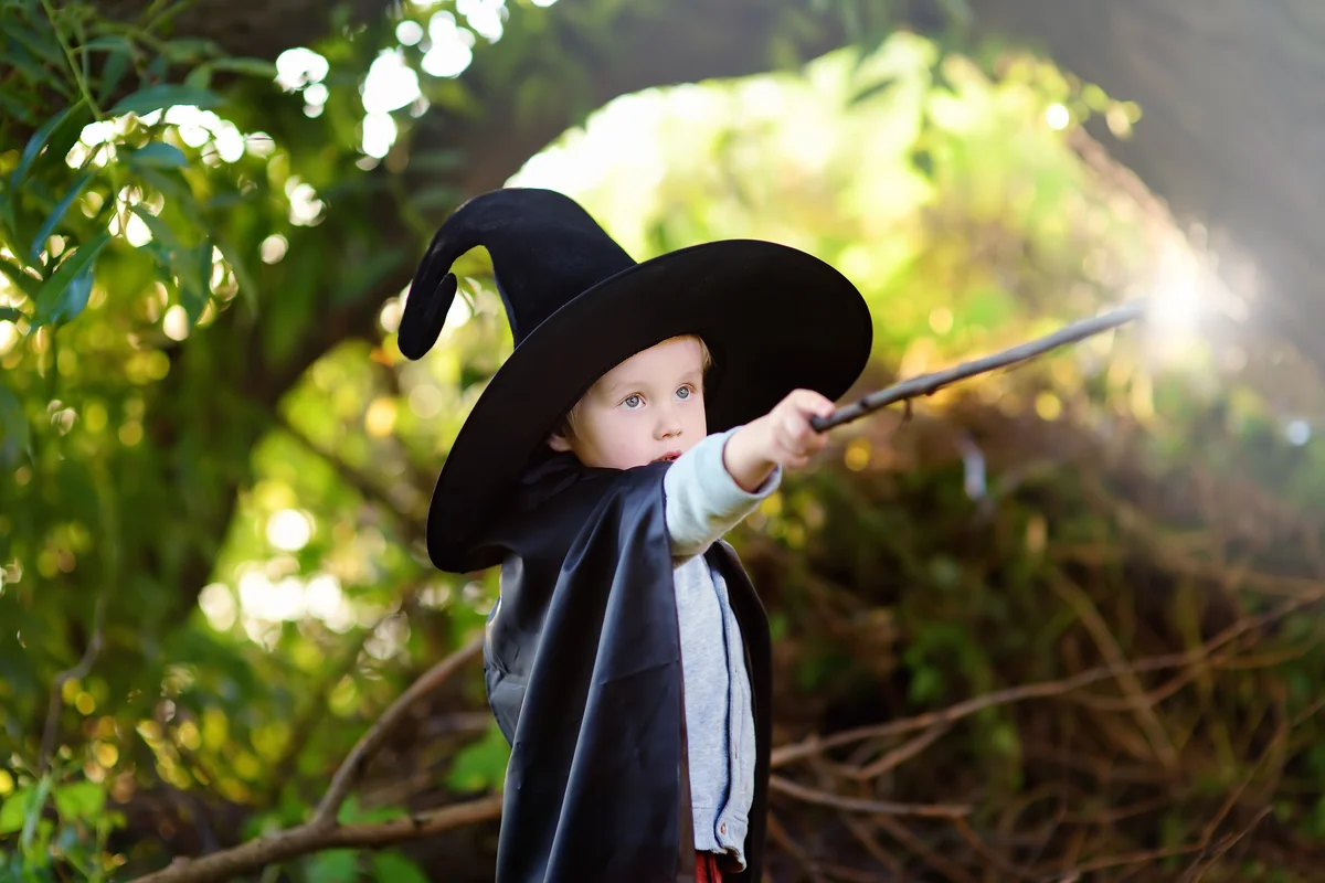 Little boy in pointed hat and black cloak playing with magic wand outdoors