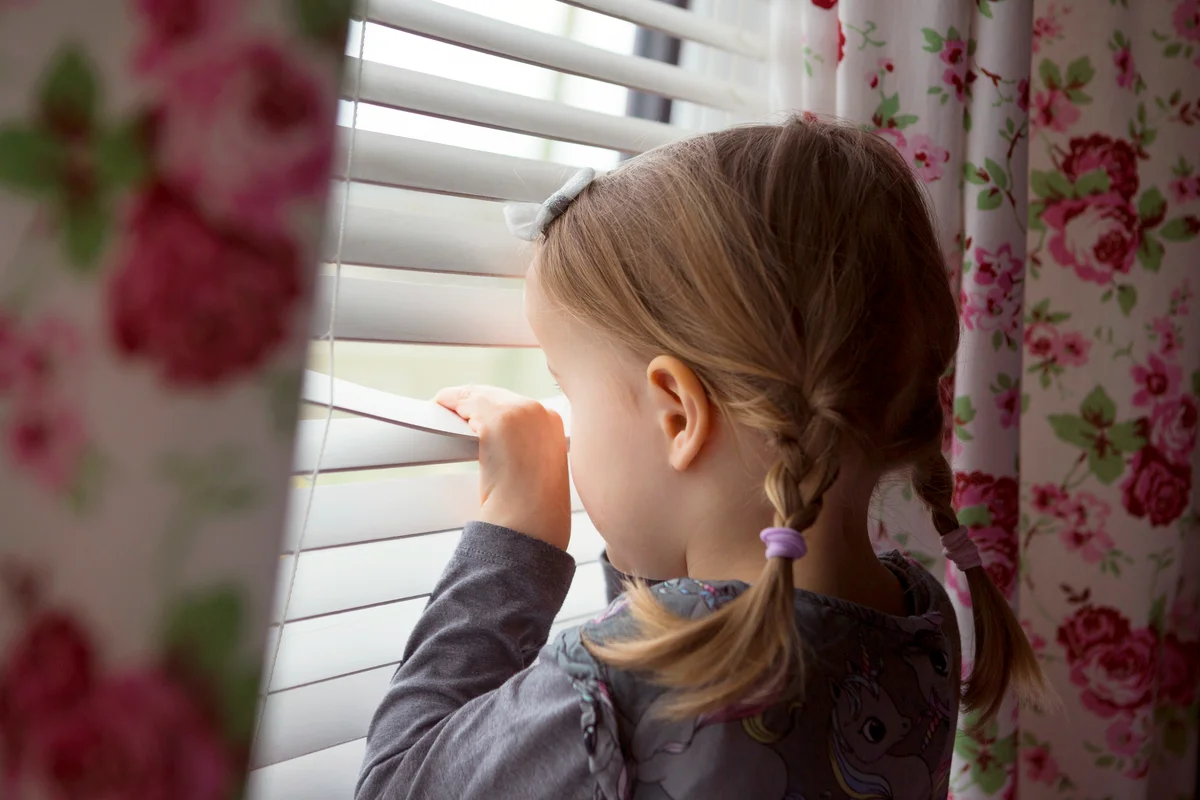 Child indoors peering out through window blinds