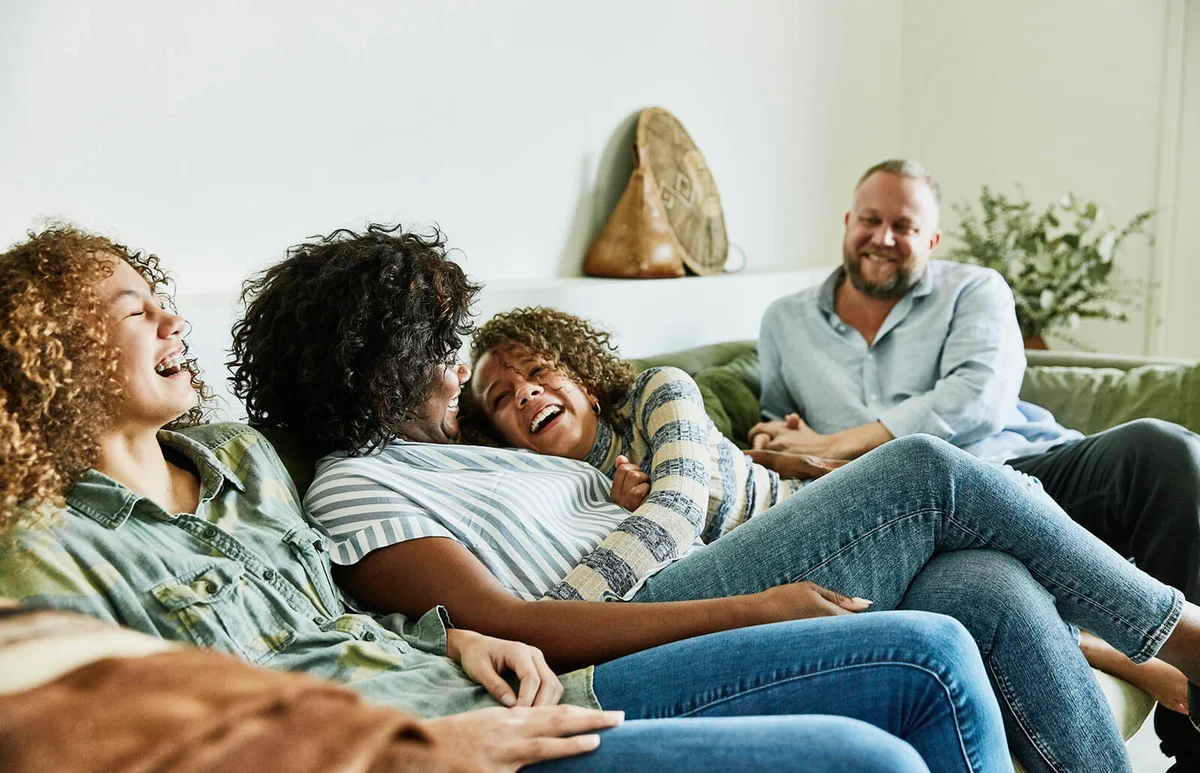 Laughing daughter embracing mother while sitting on couch with family in living room