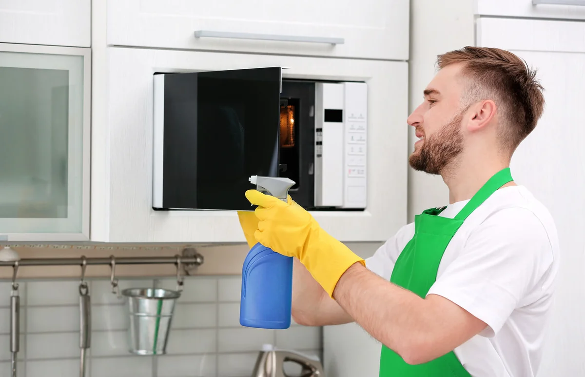 Young man in uniform cleaning microwave oven in kitchen