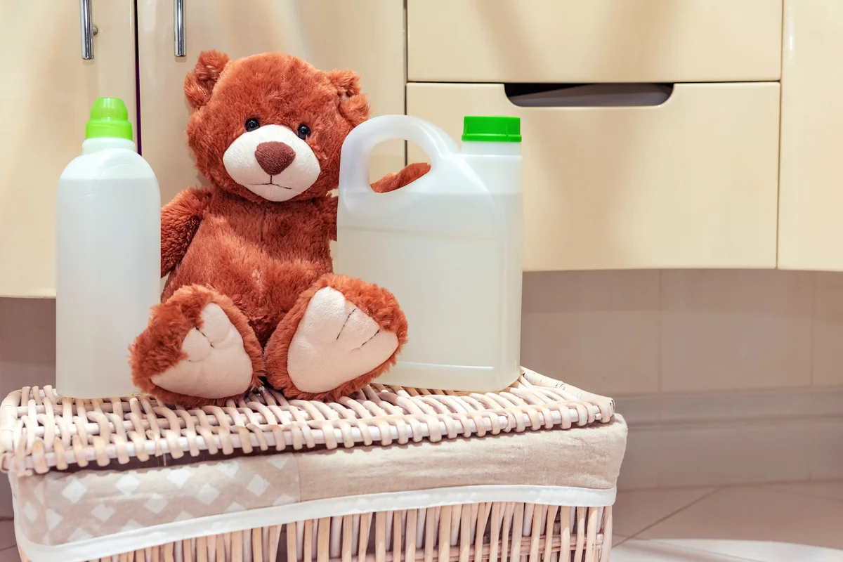 A stuffed toy teddy bear is sitting on laundry basket in the bathroom next to detergent and rinse aid