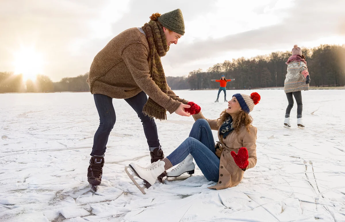 Man helping ice skating woman up on icy surface of frozen lake