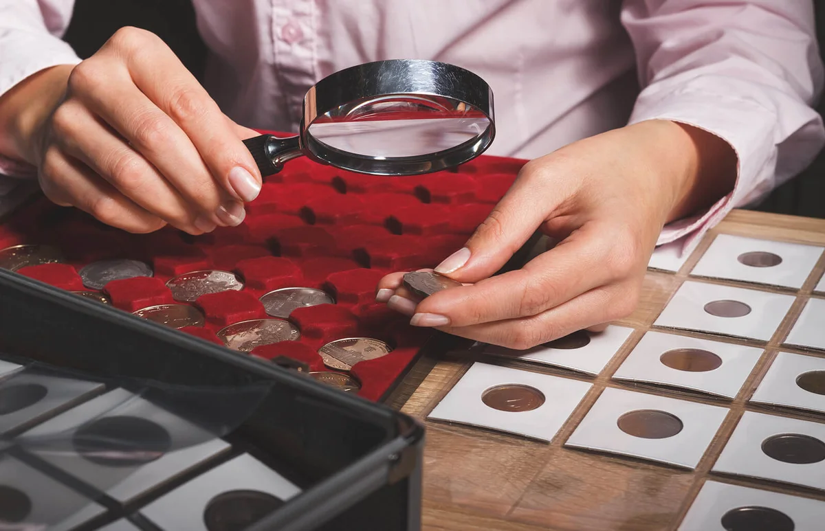 Box with collectible coins in the cells and a hand with coin through the magnifying glass