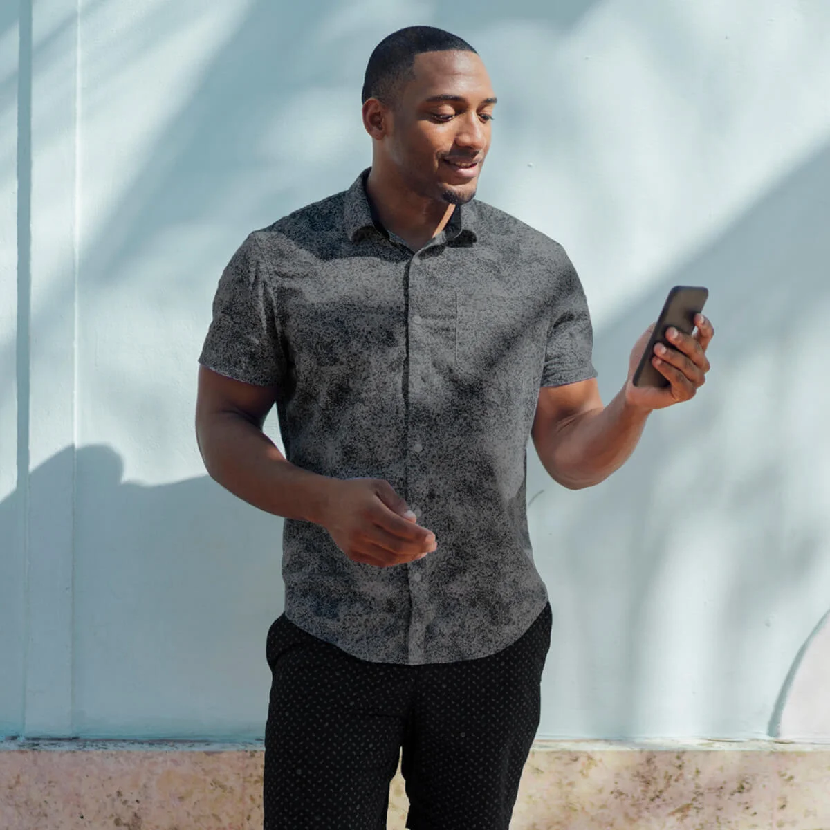 Smiling young man wearing shirt looking at cell phone at a wall