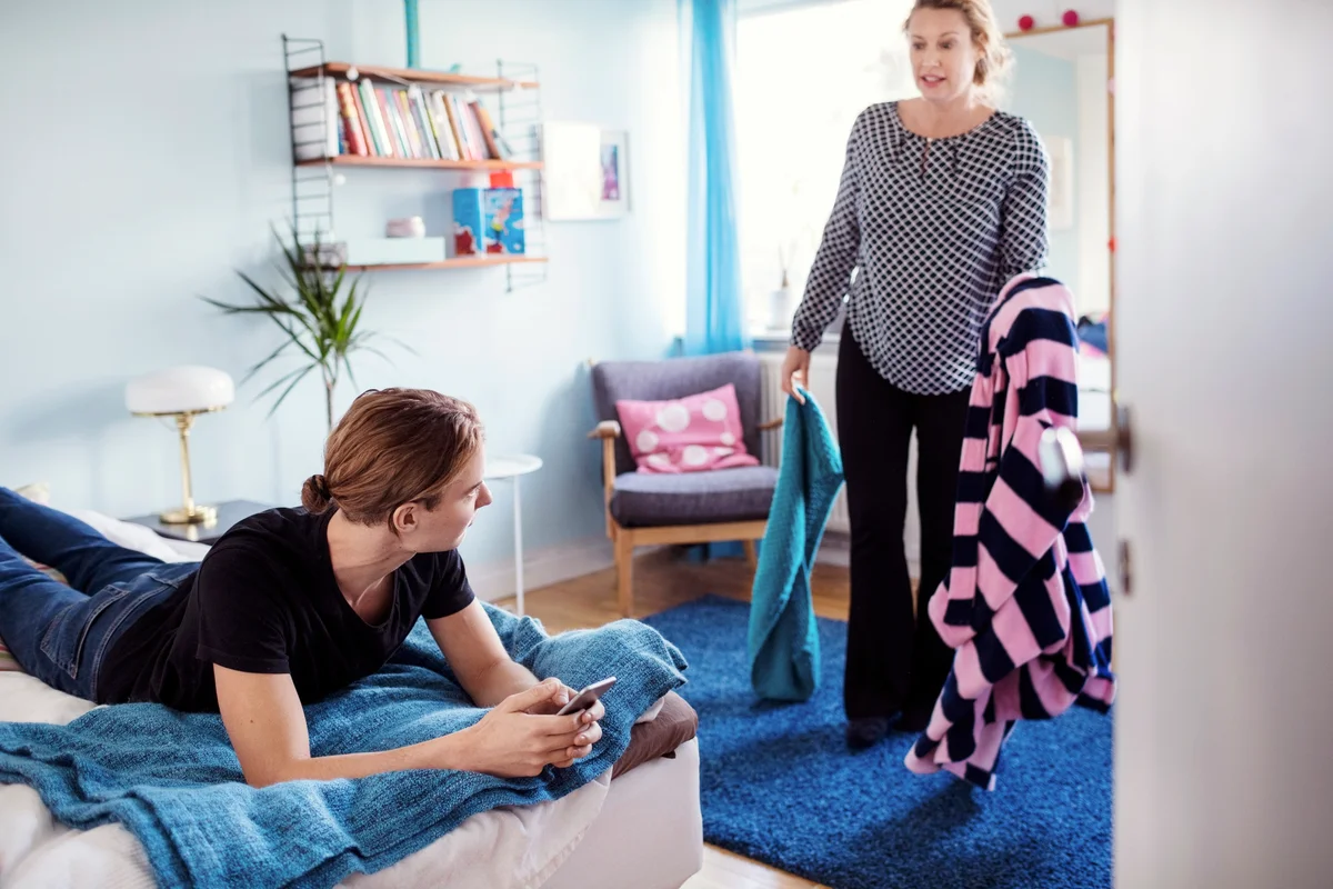 Angry woman holding clothes while looking at son lying on bed
