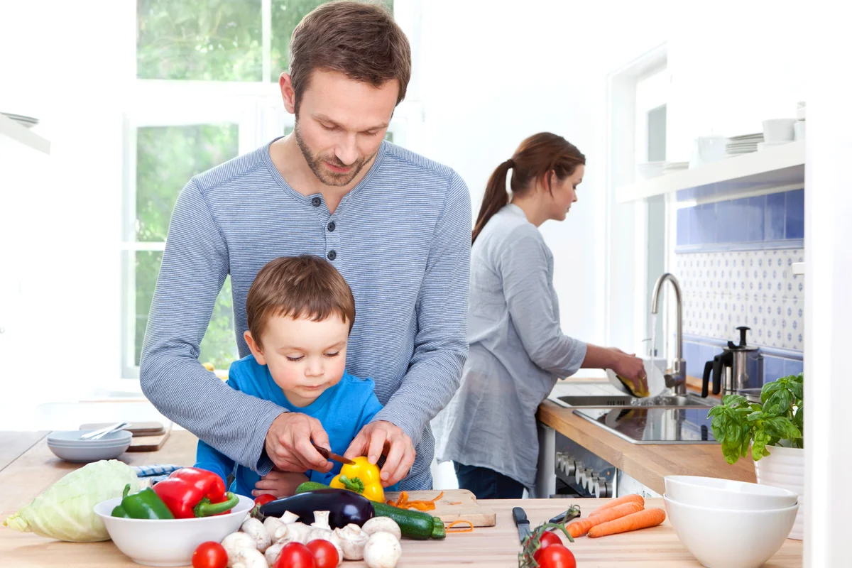 Father and son chopping vegetables and mother washing dishes in background