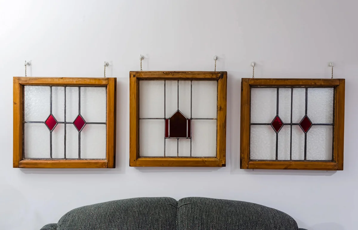 three Wooden framed English stained glass windows with red glass patterns hanging on a white wall above a green couch