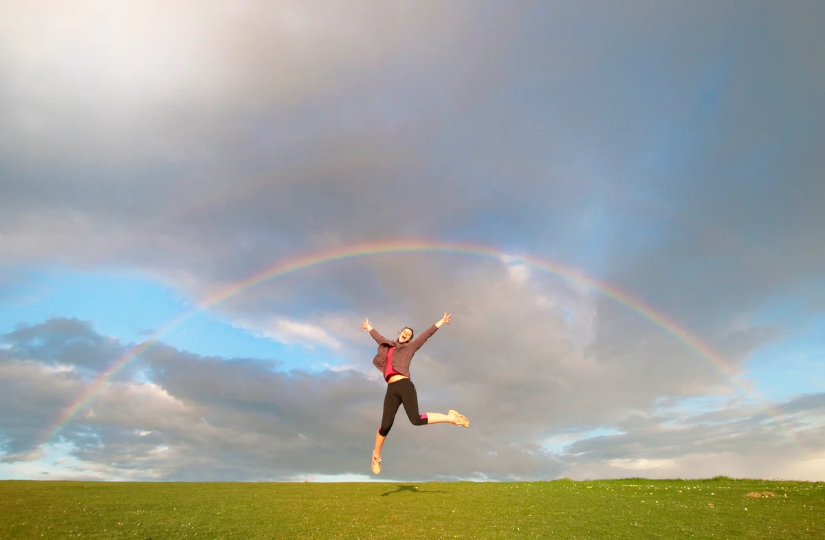 woman jumping in air celebrating feng shui good luck underneath a rainbos in the sky