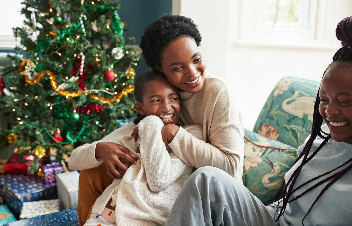 Smiling young woman embracing girl while sitting on sofa at home during Christmas festival