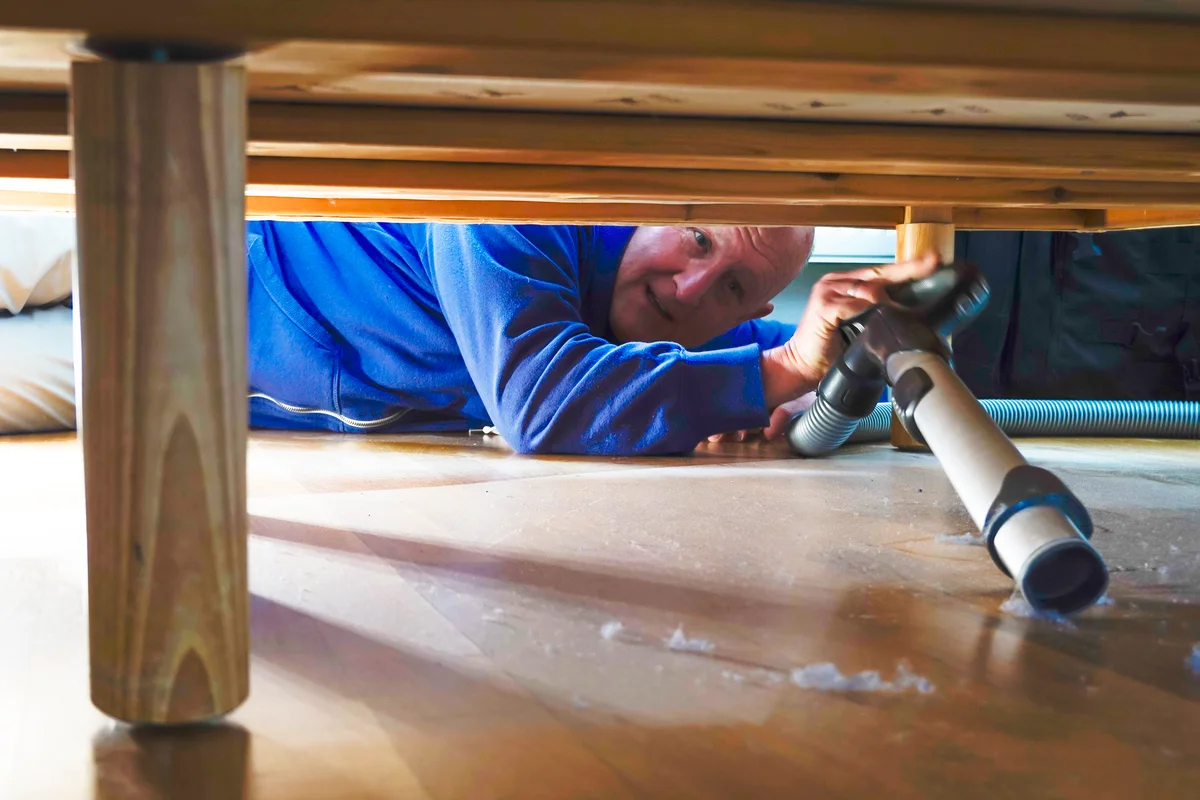A man vacuums dust under a bed in an apartment