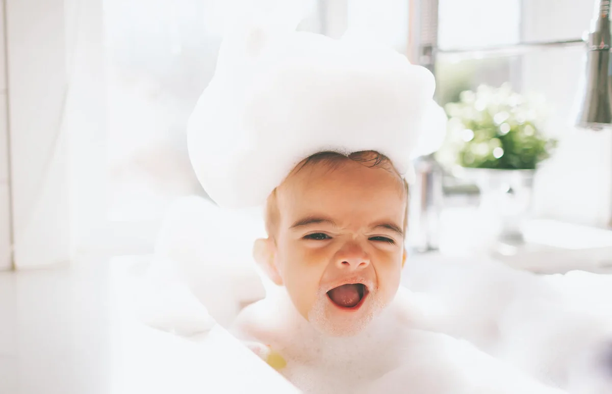 Cute Happy Baby Boy Taking Bubble Bath In Kitchen Sink At Home