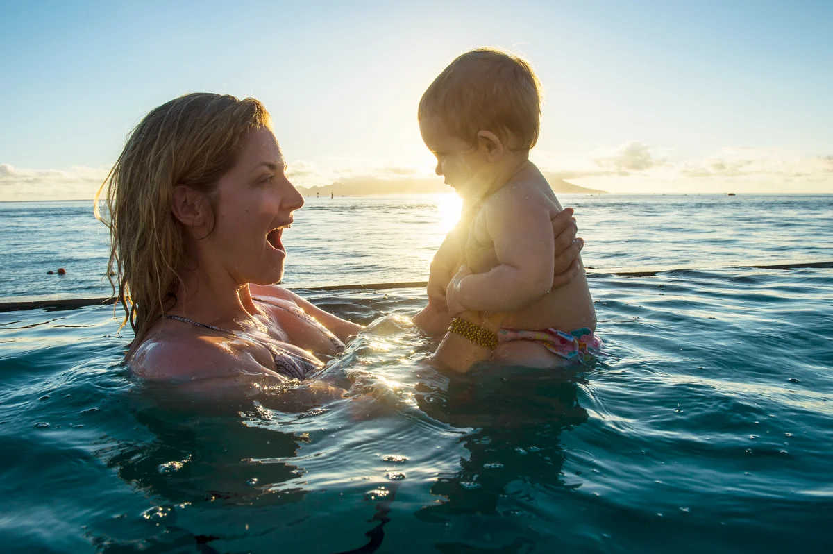 Woman playing with her little baby in the ocean at sunset