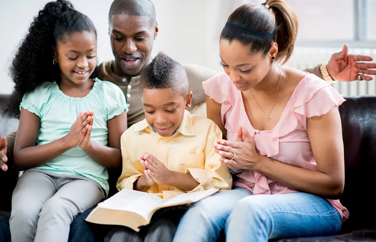 family sitting on their sofa together, folding their hands for prayer with the bible open