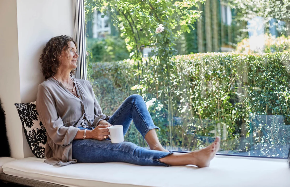 Mature woman sitting at the window at home with cup of coffee