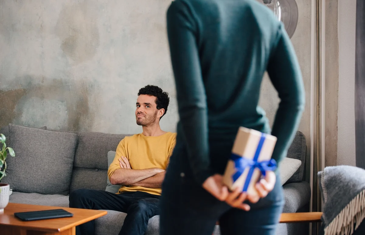 man looking at girlfriend hiding gift box while standing in living room