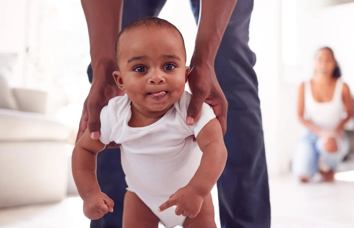 Parents Encouraging Smiling Baby Daughter To Take First Steps And