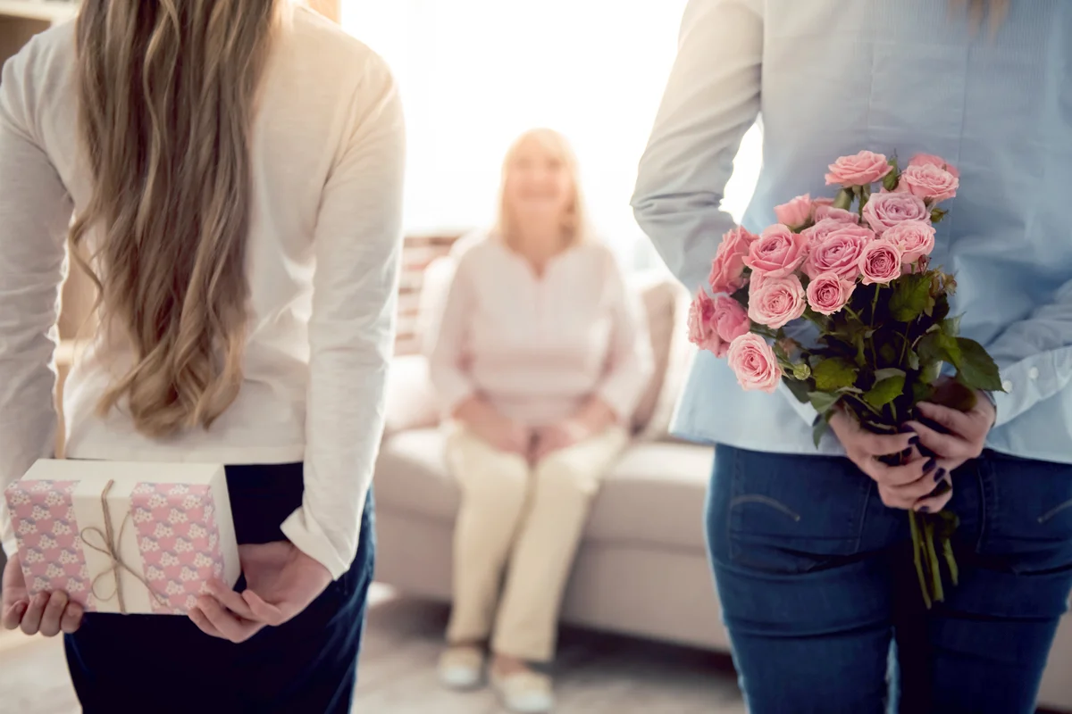 Teenage girl and older daughter are hiding flowers and a gift box for their mom