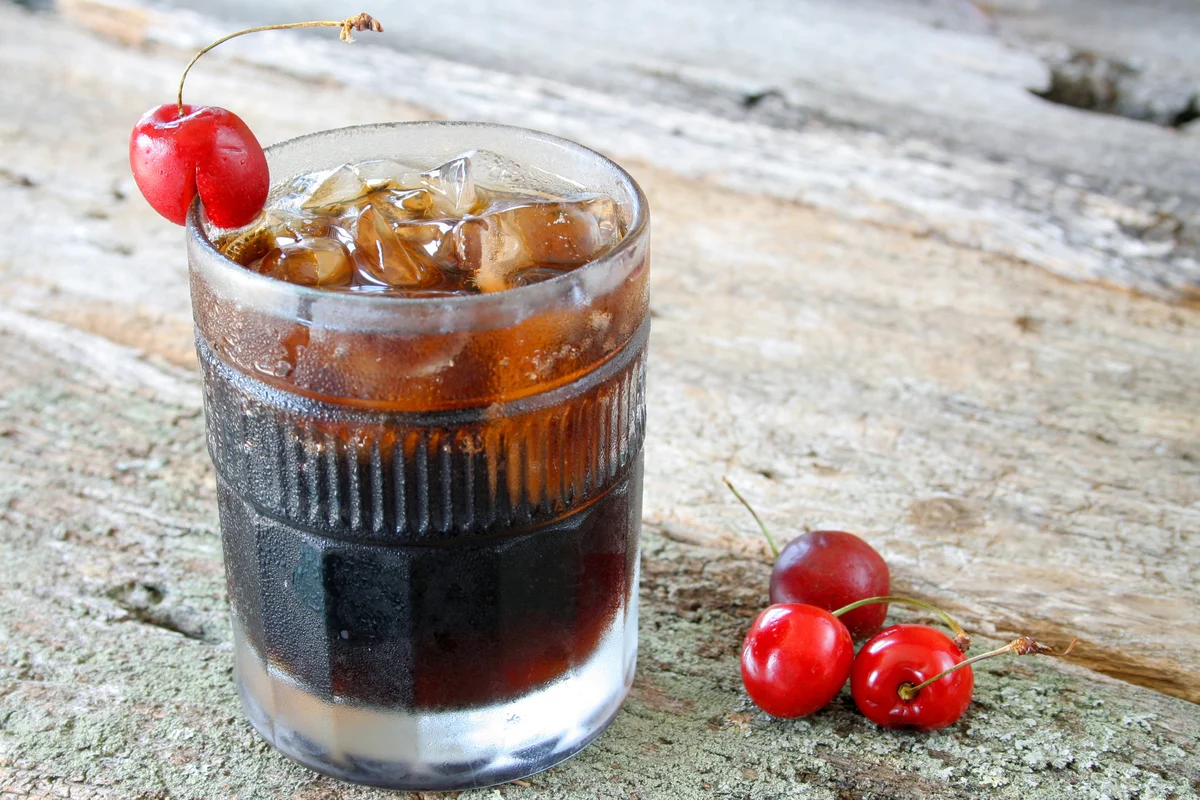 A glass of joe collins and ice cubes with fresh ripe cherries as a garnish