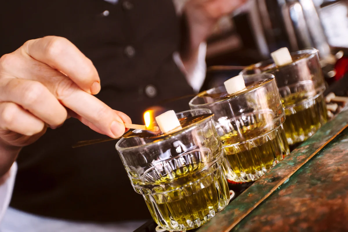Human hand holding light match warming sugar cubes over spoon on absinthe glasses at the bar