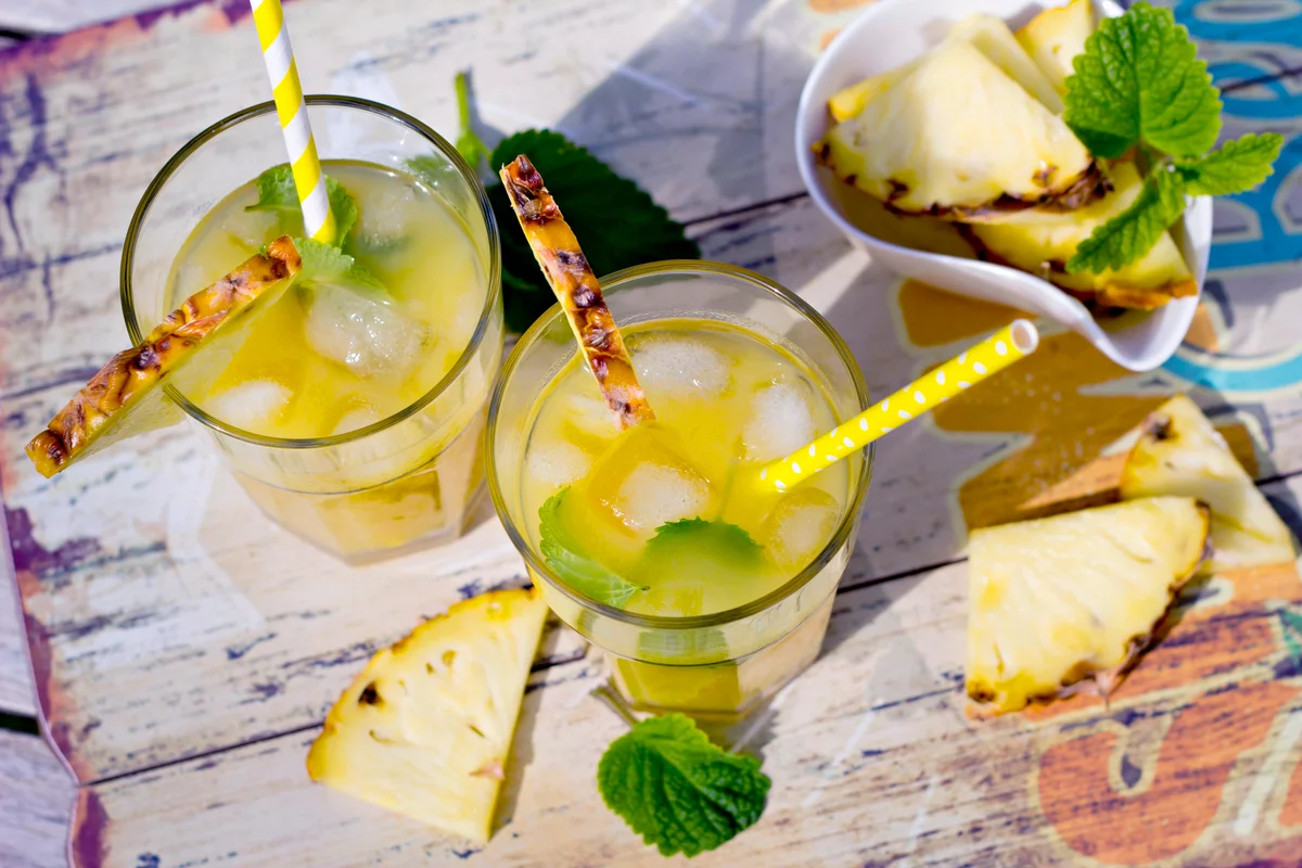 Two glasses with ice, cocos sirup and ananas juice, garnished with pieces of ananas and lemon balm