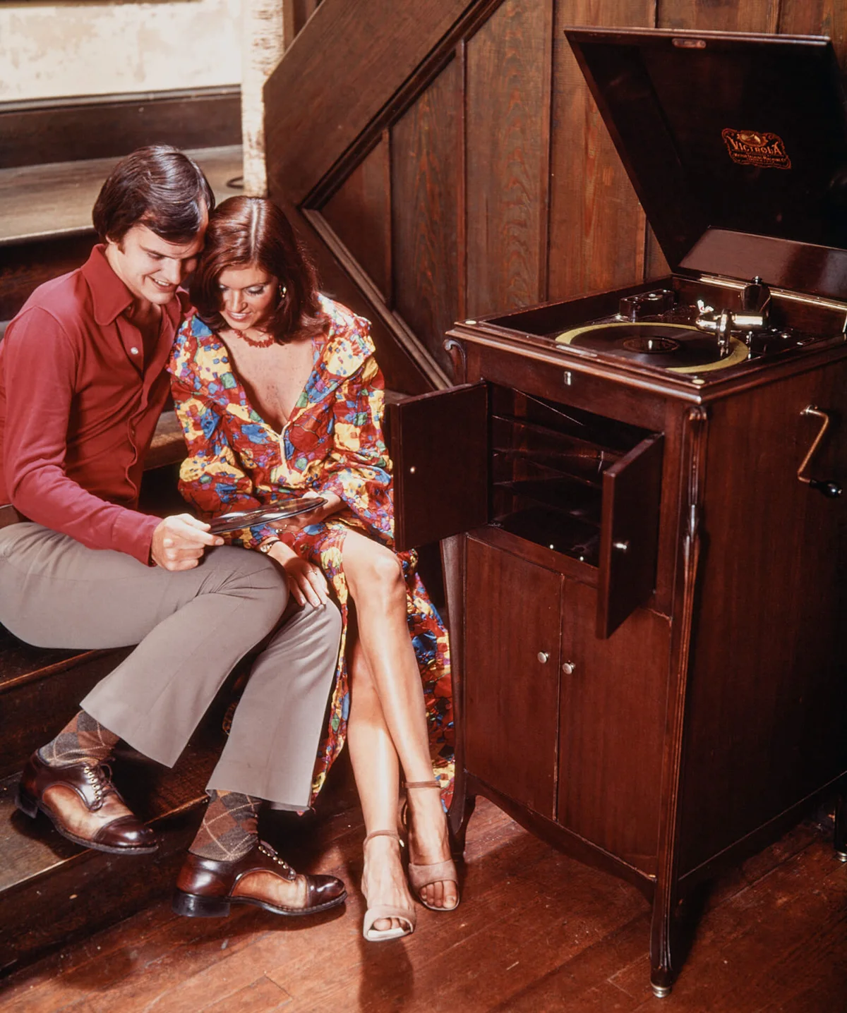 1970s COUPLE SITTING CLOSE TOGETHER LISTENING TO ANTIQUE WINDUP SPRING DRIVEN AUDIO VICTROLA RECORD PLAYER | Getty Editorial Use